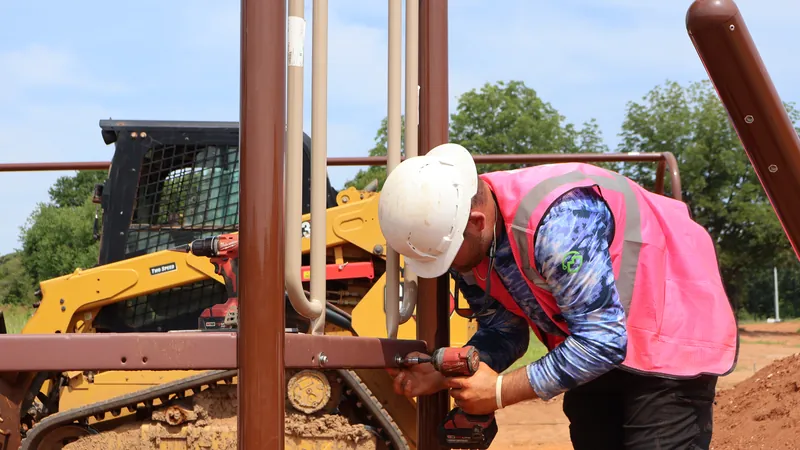An installer securing playground post hardware with a power drill, a skid steer staged behind.