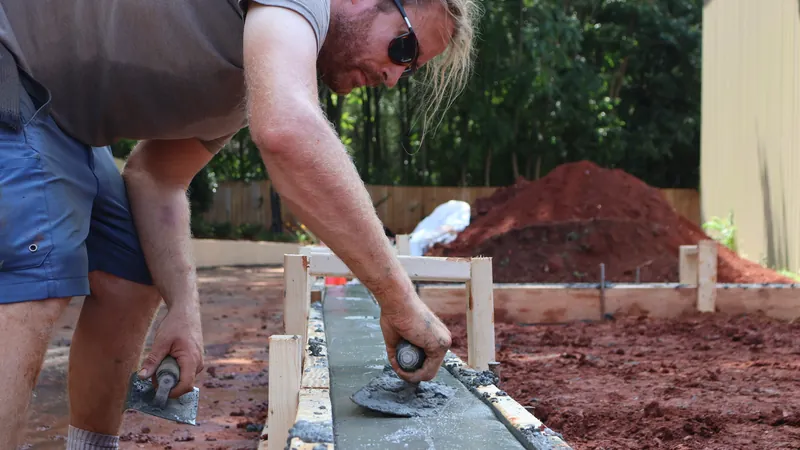 A crew member hand-troweling a concrete pour set within wood forming boards on a graded site.